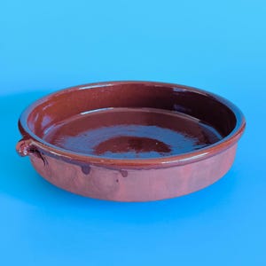 May include: A round, shallow terracotta baking dish with a glossy, dark brown glazed interior and a matte, reddish-brown exterior. The dish features a small, looped handle on one side and a smooth, circular rim, showcasing a classic earthenware design.