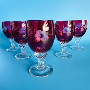 May include: A set of six ruby red wine glasses. Each glass has a clear, twisted stem and a floral design etched on the bowl. The glasses are displayed on a blue background, highlighting their elegant design.