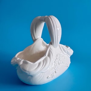 May include: A white ceramic basket featuring a high, arched handle and a sculpted, flowing rim. The exterior surface is decorated with embossed floral patterns and draped fabric-like textures, creating a detailed, three-dimensional appearance.