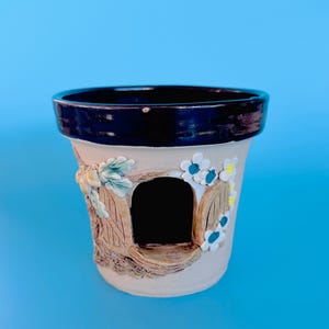 May include: A ceramic plant pot with a fairy house design. The pot has a black rim, a beige body, and a detailed facade with a door and floral accents in blue and yellow. Suitable for small plants, it is a charming decorative item.