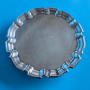 May include: A circular silver tray with a decorative, scalloped edge. The tray has a reflective surface and is set against a bright blue background. The tray appears to be made of metal and has a subtle inscription.