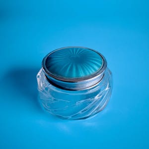 May include: A clear glass jar with a silver rim and a teal lid. The lid has a radial design. The jar has a spiral design on the glass. The jar is on a blue background.