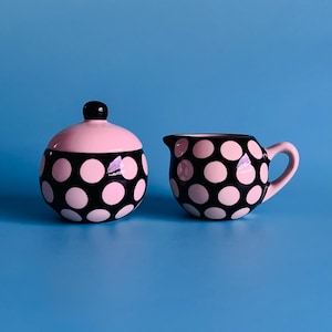 May include: A pink and black polka dot sugar bowl with a lid and a matching creamer. The sugar bowl has a black knob on the lid. The creamer has a handle and a spout. Both items are on a blue background.