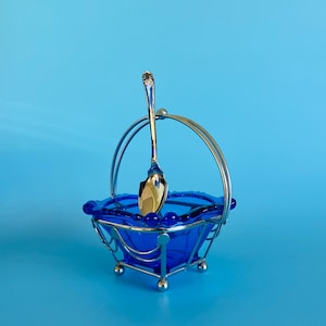 May include: A vibrant cobalt blue glass dish with a silver metal handle and base. A silver spoon is placed inside. The dish is square with a beaded rim, set against a blue backdrop.