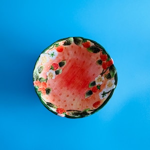 May include: A ceramic bowl with a strawberry design. The bowl is pink with green leaves and red strawberries.
