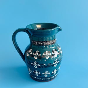 May include: A teal ceramic jug featuring a rounded body, a curved handle, and a pouring spout. The exterior is decorated with horizontal bands of hand-painted white and dark grey floral patterns. A small rectangular label is visible inside the rim.