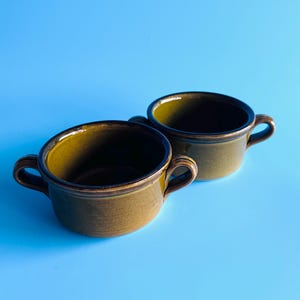 May include: Two matching ceramic soup bowls in a deep olive green glaze. Each bowl features a rounded body, a smooth interior, and two small, curved handles on opposite sides. The exterior finish is consistent with a glossy, earthy tone.