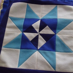 May include: A square, quilted pillow with a starburst design. The star is composed of white, light blue, and dark blue triangles. The pillow has a dark blue border and a ruffled edge.