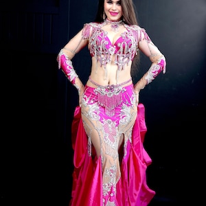 May include: A woman wearing a pink and silver belly dancing costume with a long skirt and intricate embellishments. The costume features a bra top, a belt, and a skirt with a slit up the front. The woman is smiling and looking at the camera.