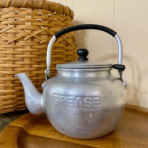 May include: A vintage aluminum teapot with a black handle and lid knob. The word "GREASE" is embossed on the side. The teapot is sitting on a wooden tray, with a woven basket in the background.
