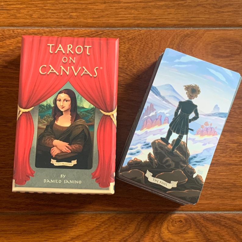 May include: Tarot On Canvas deck box with a red velvet curtain design and a depiction of the Mona Lisa. The box is next to a single card from the deck, The Fool, which features a person standing on a rock with a blue and white landscape in the background.
