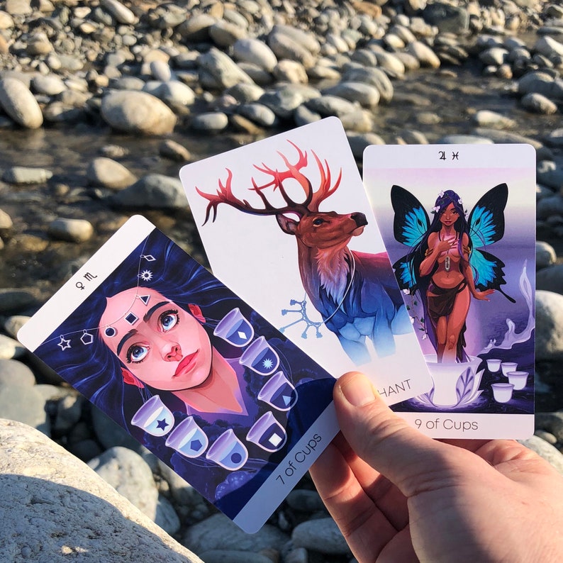 May include: Three tarot cards, the 7 of Cups, the 9 of Cups, and the 2 of Wands. The 7 of Cups card features a woman with long blue hair and a crown of stars, surrounded by seven cups with different symbols. The 9 of Cups card features a woman with long black hair and a blue dress, holding a cup. The 2 of Wands card features a deer with antlers and a blue and white collar.