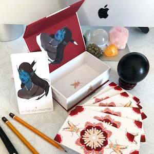 May include: A set of tarot cards featuring a blue and white illustration of a woman with a crown on her head. The card reads "II - The High Priestess". The cards are in a box with a red lid and a white interior. The box is open and the cards are fanned out.