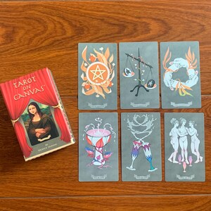 May include: Tarot on Canvas deck featuring illustrations of the Ace of Pentacles, Two of Swords, Three of Wands, Ace of Cups, Two of Cups, and Three of Cups. The cards are gray with colorful illustrations.