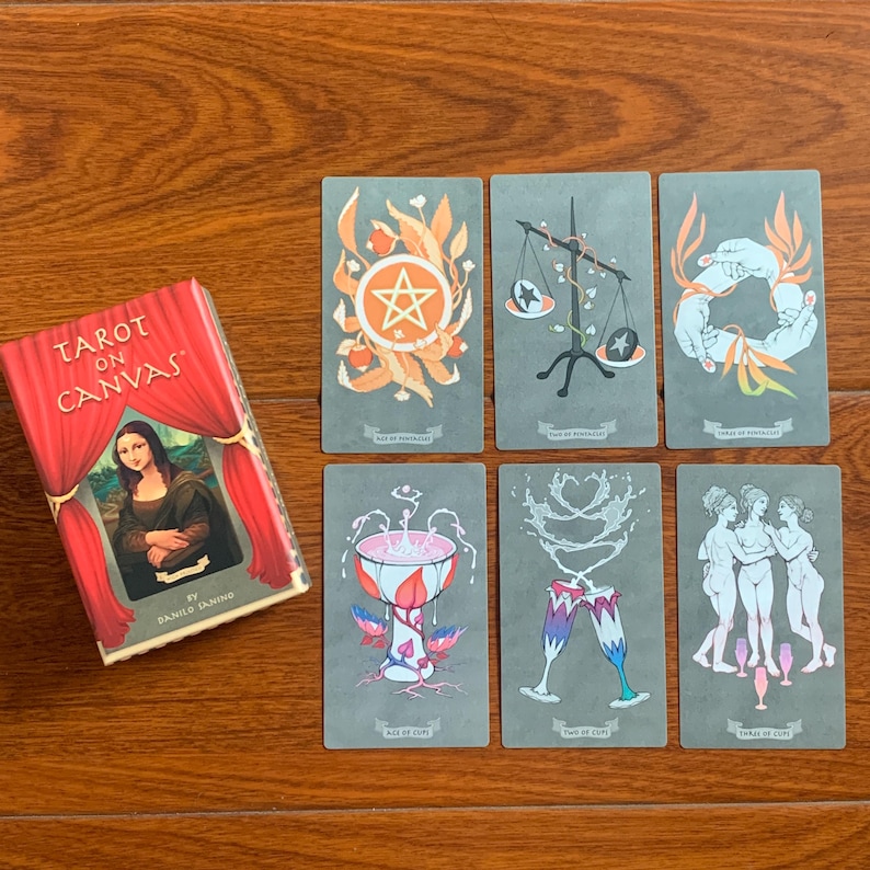 May include: Tarot on Canvas deck featuring illustrations of the Ace of Pentacles, Two of Swords, Three of Wands, Ace of Cups, Two of Cups, and Three of Cups. The cards are gray with colorful illustrations.