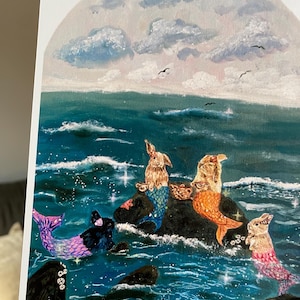 May include: An art print depicting rabbit mermaids in a seascape. The rabbits have colourful mermaid tails and are on rocks in the ocean. The sky is filled with clouds and a few birds are flying above the sea.