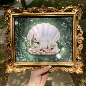 May include: A white unicorn lying inside a large, pink seashell with a green leafy background. The unicorn has a gold horn and is surrounded by bubbles.