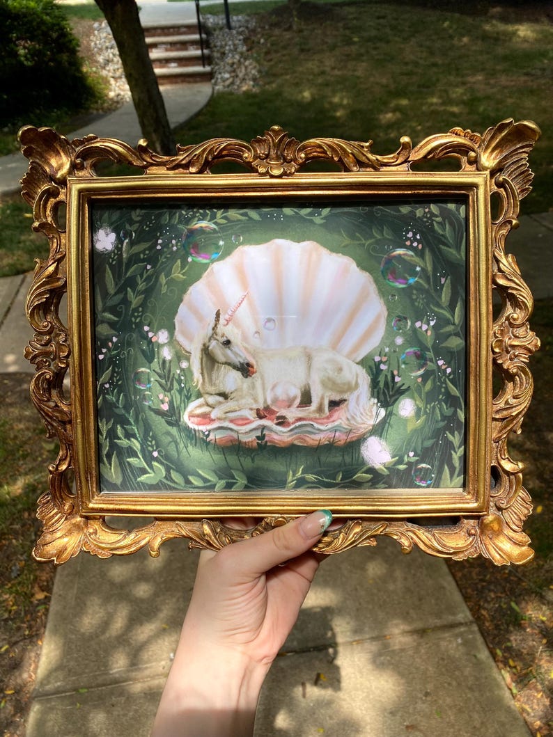 May include: A white unicorn lying inside a large, pink seashell with a green leafy background. The unicorn has a gold horn and is surrounded by bubbles.