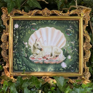 May include: A white unicorn with a pink horn is lying down inside a large, pearl-coloured seashell. The seashell is surrounded by green leaves and small, iridescent bubbles. The image is framed in a gold ornate frame.