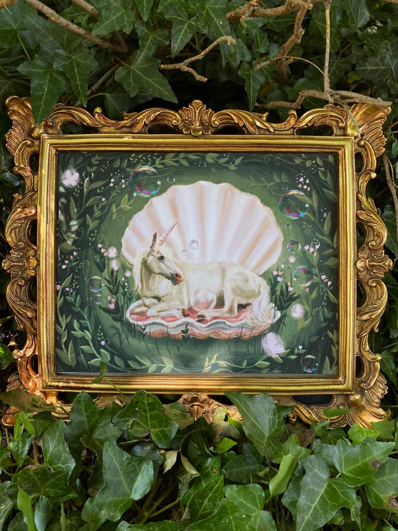 May include: A white unicorn with a pink horn is lying down inside a large, pearl-coloured seashell. The seashell is surrounded by green leaves and small, iridescent bubbles. The image is framed in a gold ornate frame.