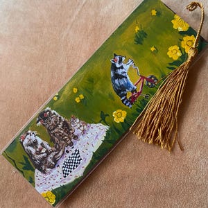 May include: Hand-painted bookmark featuring a raccoon riding a red bicycle on a green field with yellow flowers. The bookmark also depicts cats on a white and pink blanket with a black and white checkered pattern. A gold tassel hangs from the top.