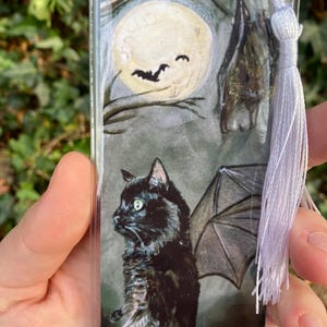 May include: A black cat with wings stands in front of a full moon. The cat has green eyes and a black and white striped tail. The moon is surrounded by dark branches. A bat hangs upside down from a branch above the cat.