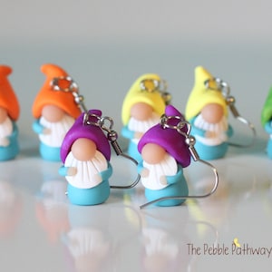 May include: A set of six colorful gnome earrings. Each earring features a gnome with a different colored hat and a white beard. The gnomes are wearing blue shirts and have a white shell in their hands. The earrings are made of polymer clay and have silver hooks.
