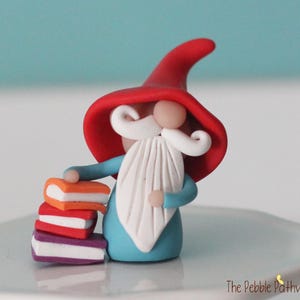 May include: A small clay figurine of a gnome wearing a large red pointed hat, a blue tunic, and a long white beard and mustache. The gnome stands next to a stack of three miniature books colored orange, red, and purple with white pages.