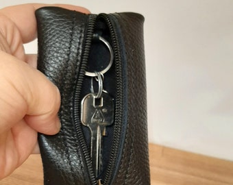 May include: A black leather key holder with a zipper closure and a silver key ring. The key holder is open and a silver key is visible inside.