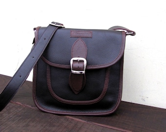 Può includere: Una borsa a tracolla in pelle marrone con una patta nera e una fibbia argentata. La borsa ha una tracolla in pelle marrone e un bordo in pelle marrone.