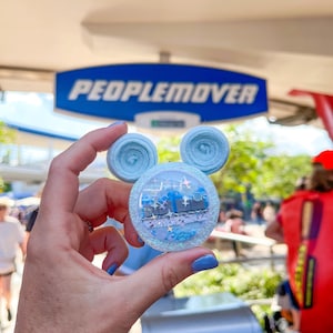 May include: A light blue Mickey Mouse-shaped accessory with a circular center featuring a train design, blue and silver glitter, and a blue bow. The sign in the background reads "PEOPLEMOVER".