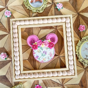 May include: A decorative wooden frame with a beaded border surrounds a pink mouse-shaped design adorned with a floral arrangement. The mouse has pink ears and a floral fabric center. Small pink roses and gold framed miniature paintings are scattered around the frame.