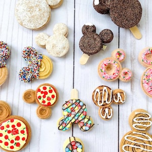 May include: A variety of Mickey Mouse shaped cookies decorated with sprinkles, chocolate, and icing. The cookies are arranged on a white wooden surface.