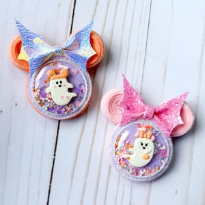 May include: Two circular hair clips featuring ghost-shaped charms inside clear shaker domes. The left clip has an iridescent bow and orange felt ears, while the right clip has a pink glitter bow and light pink felt ears, both filled with colorful confetti.