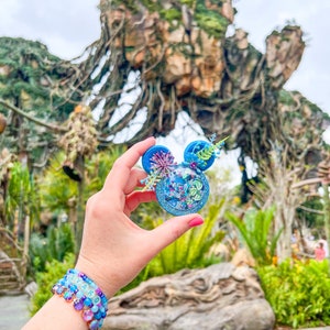 May include: A blue and green Mickey Mouse ear headband with a floral design and glitter. The headband is being held in front of a large rock formation with vines and greenery.