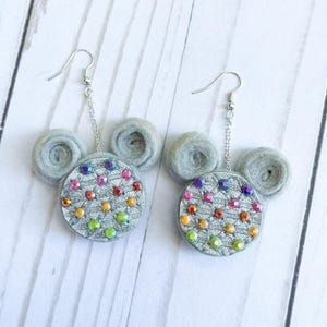 May include: A pair of handmade earrings in a mouse-ear design. The earrings have silver hooks and chains. The circular ear pieces are silver with colourful rhinestones. The mouse ears are grey.