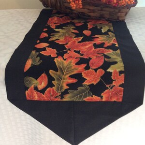 May include: A fall-themed table runner with a black border and a central panel featuring colorful autumn leaves on a black background. A woven cornucopia basket filled with orange berries and pumpkins sits on top.
