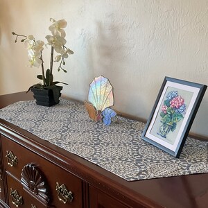 May include: A wooden dresser with a gray and white patterned table runner. A framed picture of flowers, a stained glass seashell, and a potted orchid are displayed on top. The dresser is dark brown.