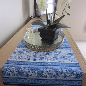 May include: A blue and white floral table runner with a decorative silver tray holding a black square planter with an orchid and a perfume bottle. The runner is on a light-colored wooden surface.