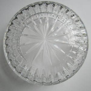 May include: A clear, round glass dish with a starburst design etched into the bottom. The rim has a textured, ridged pattern. The dish is transparent, allowing light to pass through, and sits on a white surface.