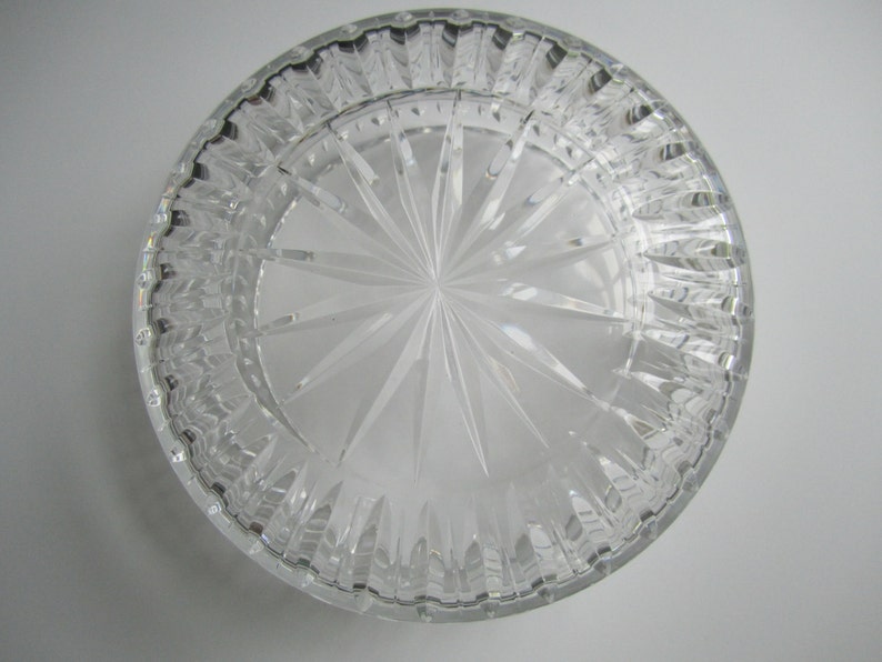 May include: A clear, round glass dish with a starburst design etched into the bottom. The rim has a textured, ridged pattern. The dish is transparent, allowing light to pass through, and sits on a white surface.