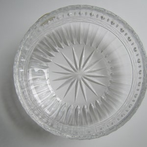 May include: Clear glass bowl with a starburst design in the center. The bowl has vertical ridges and a decorative rim with small, repeating patterns. The bowl is transparent, allowing light to pass through.