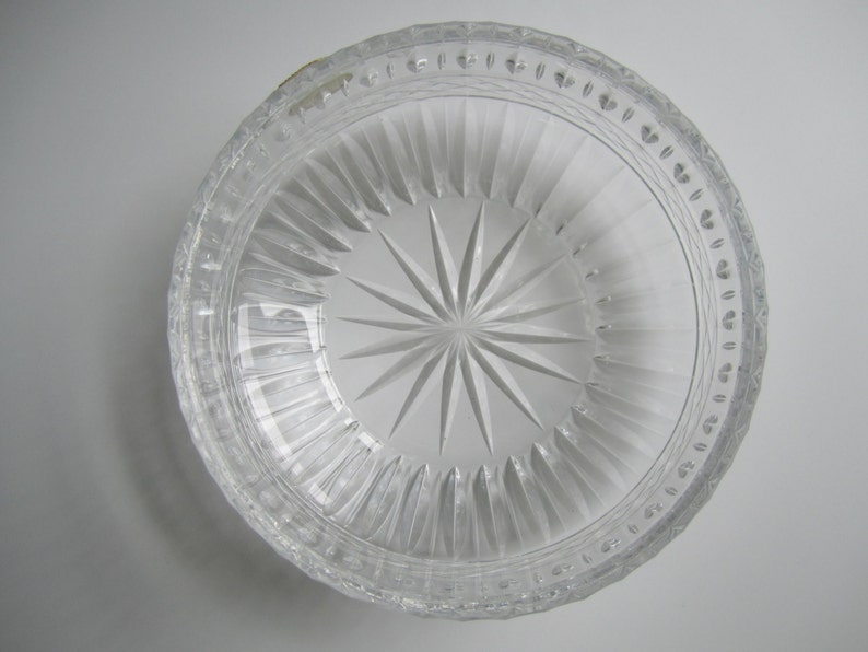 May include: Clear glass bowl with a starburst design in the center. The bowl has vertical ridges and a decorative rim with small, repeating patterns. The bowl is transparent, allowing light to pass through.