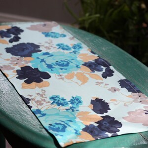 May include: A rectangular floral table runner featuring a pattern of blue, teal, and navy flowers with tan and light pink accents. The runner is displayed on a green surface.