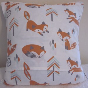 May include: Square white pillow with a repeating pattern of orange foxes, gray trees, and blue and gray geometric shapes. The foxes are in various poses. The pillow has a simple, clean design.