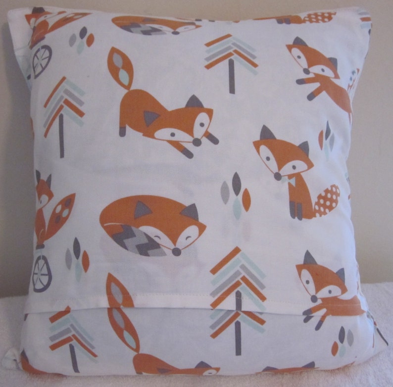 May include: Square white pillow with a repeating pattern of orange foxes, gray trees, and blue and gray geometric shapes. The foxes are in various poses. The pillow has a simple, clean design.