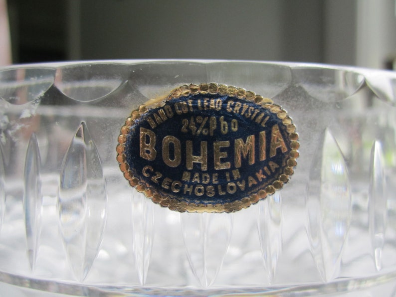 May include: Close-up of a clear crystal bowl with a decorative cut pattern. A blue and gold label reads "HAND CUT LEAD CRYSTAL 24% PbO BOHEMIA MADE IN CZECHOSLOVAKIA". The bowl's surface reflects light, highlighting its intricate design.