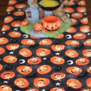 May include: A Halloween-themed table runner with a repeating pattern of orange jack-o'-lanterns, bats, stars, and moons on a dark gray background. A decorative house, cauldron, and pumpkins are placed on the runner, creating a festive display.