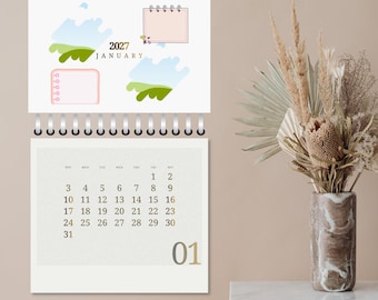 May include: A wall calendar for January 2027 featuring a spiral binding, a top section with abstract blue and green shapes and two pink note boxes, and a bottom section displaying the calendar grid with the number 01 in the bottom right corner.