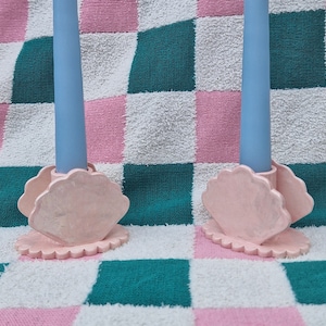 May include: Two pink seashell-shaped candle holders with light blue candles. The candle holders have a scalloped edge and are set against a pink, white, and teal checkered background.